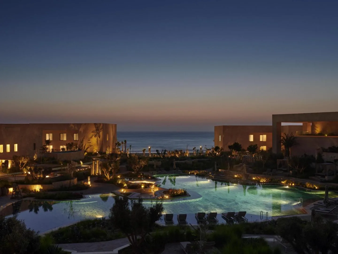 Illuminated pool at the Fairmont Taghazout Bay at dusk with the ocean behind
