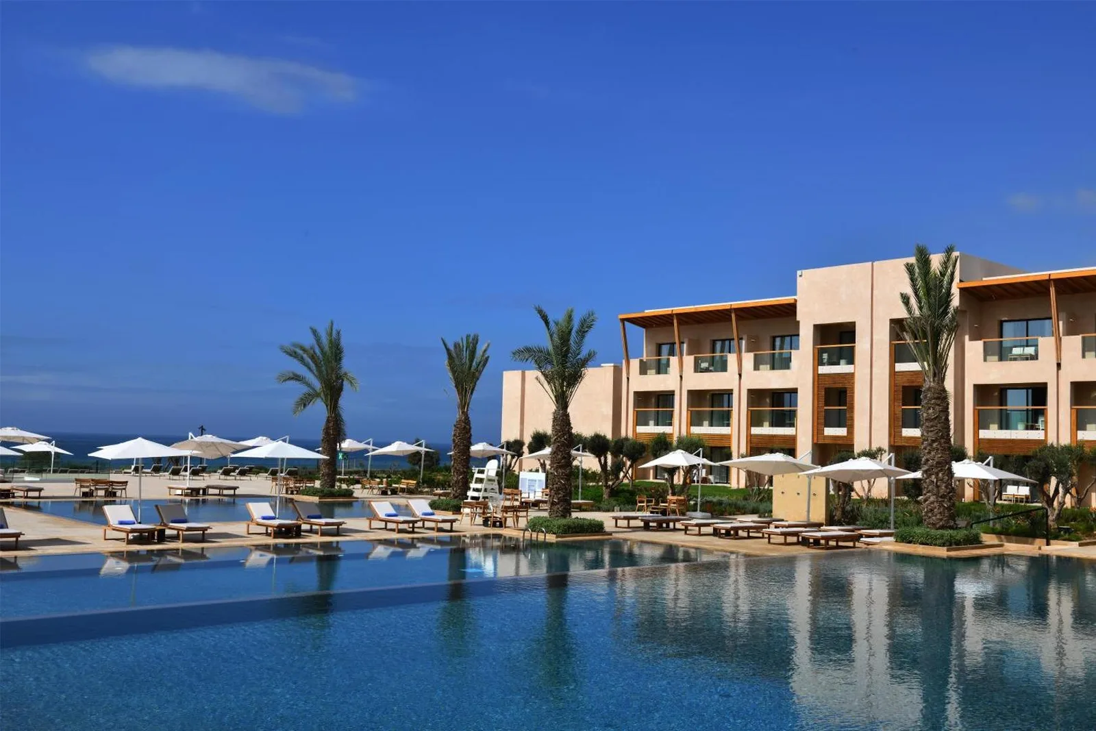 Ochre terraced building of the Hilton Taghazout Bay, with palm trees, parasols and a pool facing the ocean
