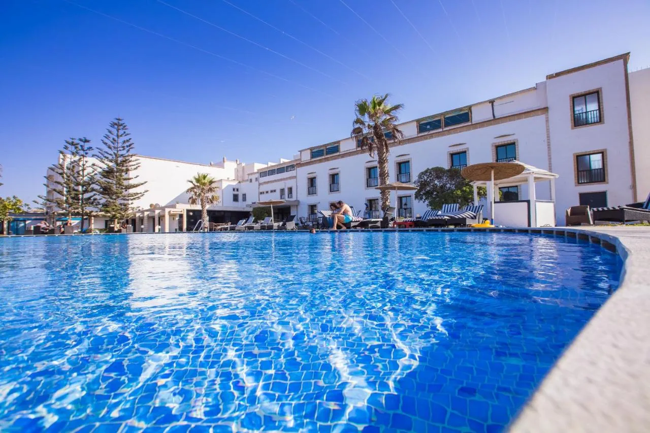 Pool at Hôtel des Îles in front of its white buildings, with palm trees and araucarias