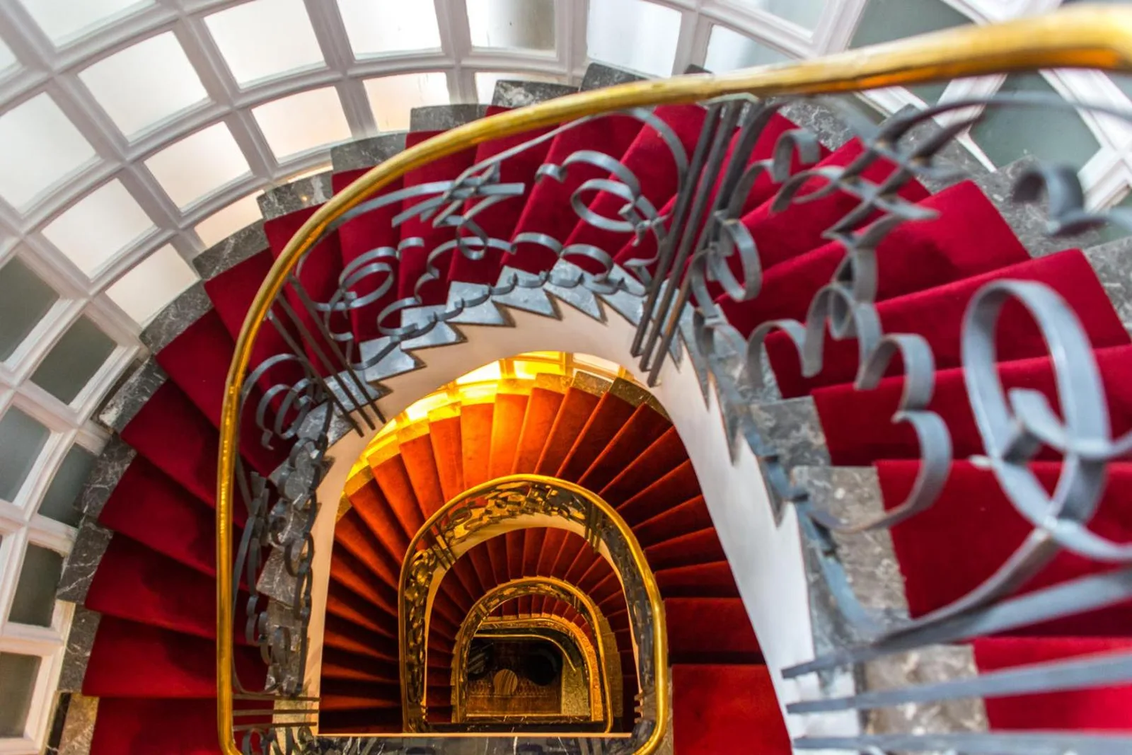 Spiral staircase with a red carpet and wrought-iron banister at the Hôtel Le Doge, seen from above under a glass roof