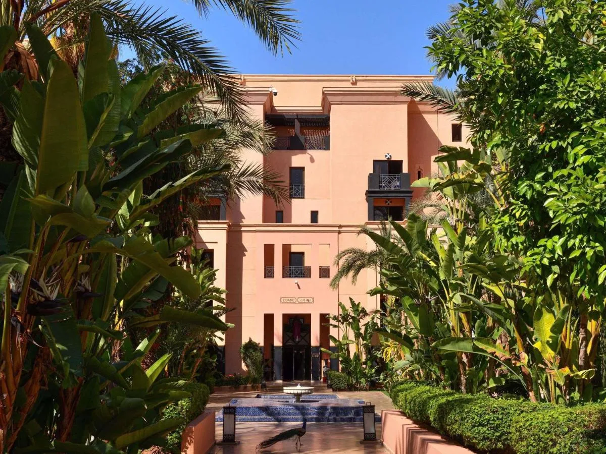 Rosa Gebäude des Mövenpick Mansour Eddahbi Marrakech, von einem tropischen Garten mit Becken aus