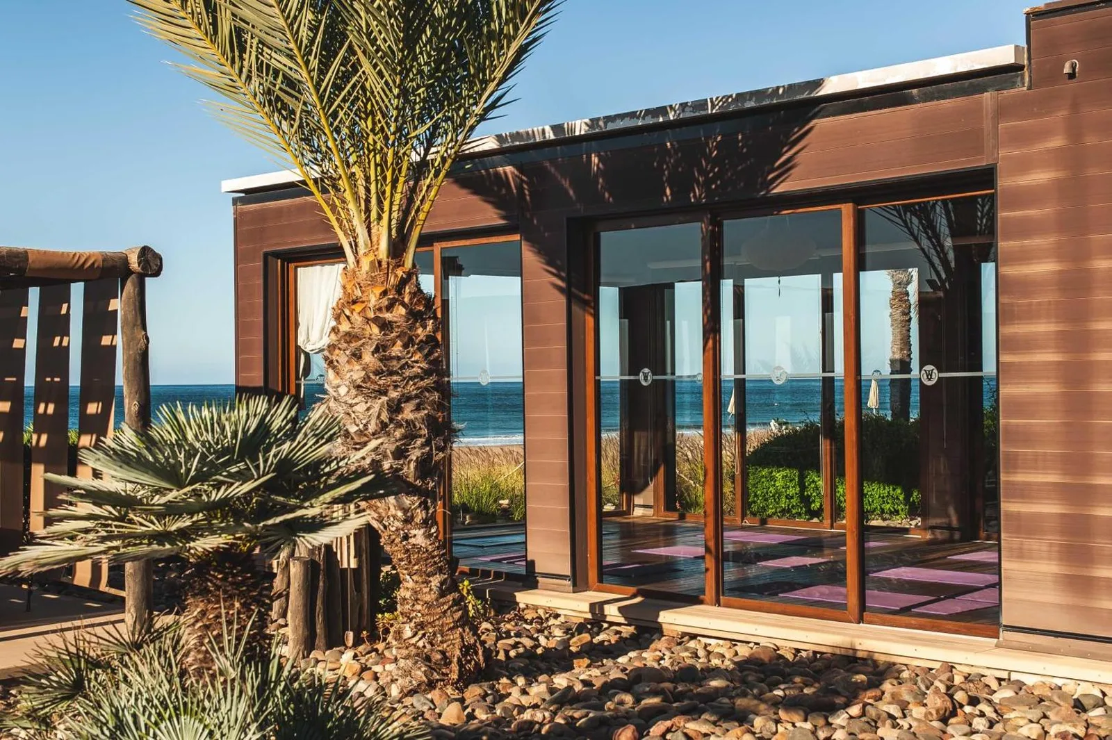 Wooden yoga pavilion at Paradis Plage with large glass doors opening onto the ocean, among palm trees and pebbles