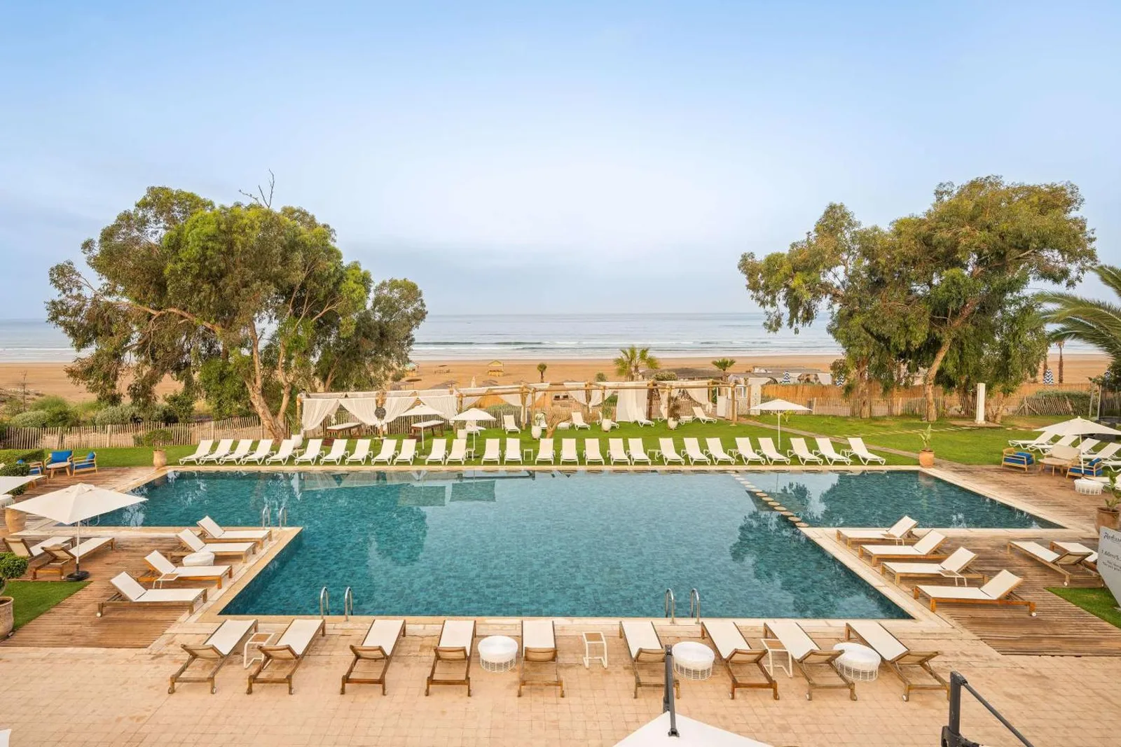 Pool at the Radisson Blu Resort Taghazout Bay Surf Village, ringed by sun loungers, with the beach and eucalyptus trees behind