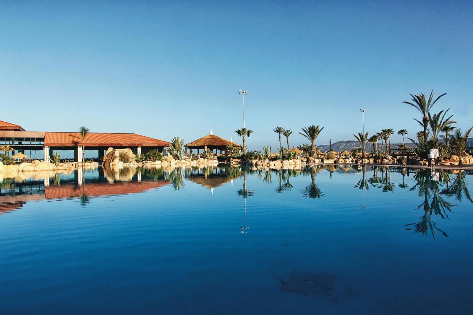 Large pool at the Riu Tikida Dunas reflecting palm trees and tiled roofs under a blue sky