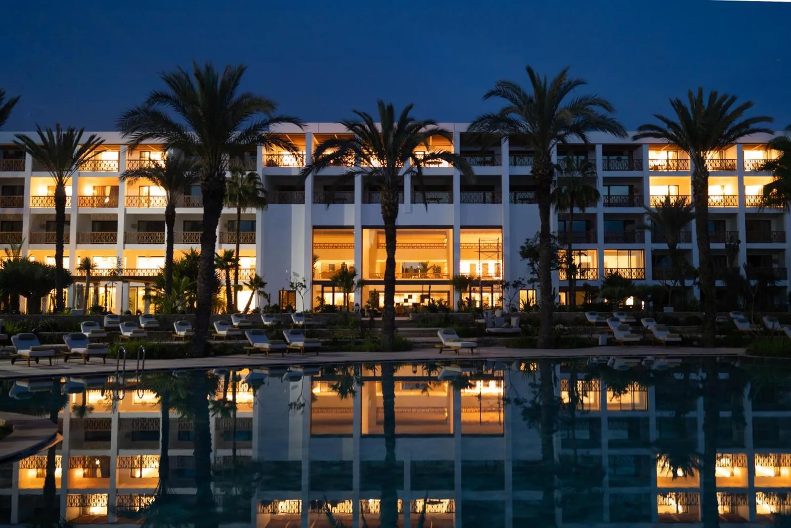 The View Agadir's illuminated façade at dusk, reflected in the pool among palm trees