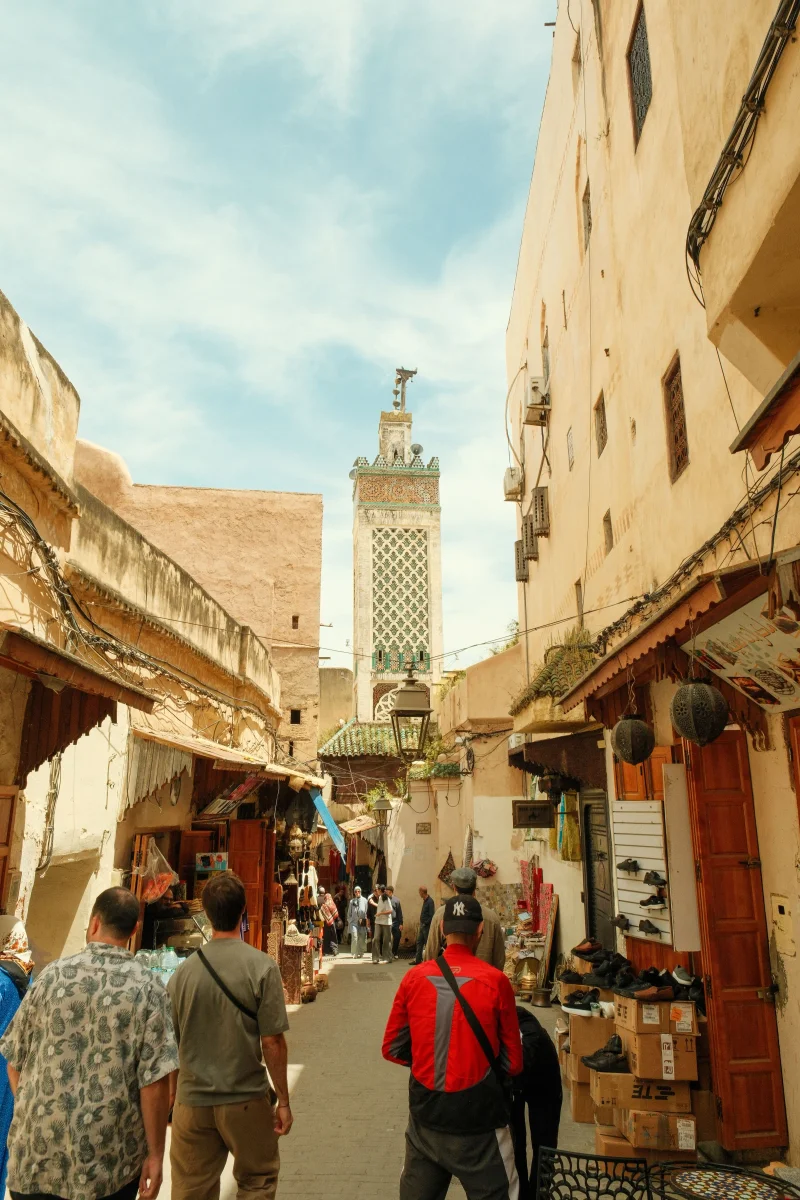Vue d'une ruelle animée dans la vieille médina de Fès, Maroc, avec le minaret de la mosquée Al Quaraouiyine se dressant entre les bâtiments traditionnels.