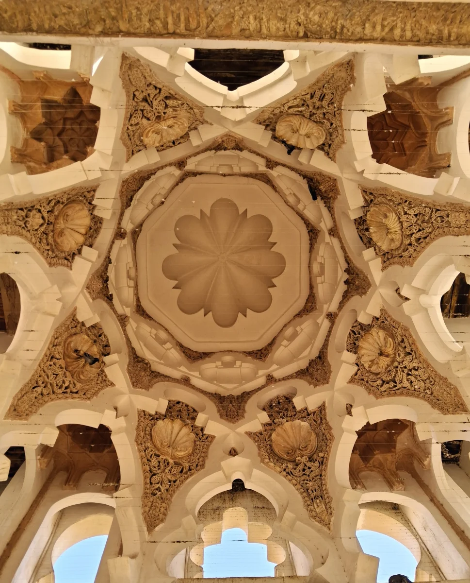 Close-up of a carved and painted ceiling with complex geometric motifs and muqarnas-like decorative elements inside the Almoravid Qubba in Marrakech.