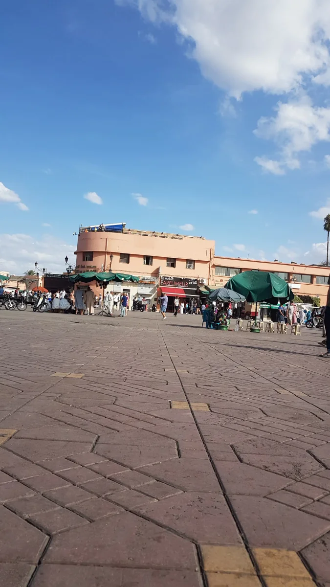 ​Vue d'une place animée, bordée de bâtiments aux tons ocres et de boutiques, avec des passants et des échoppes dans le quartier du Mellah à Marrakech.