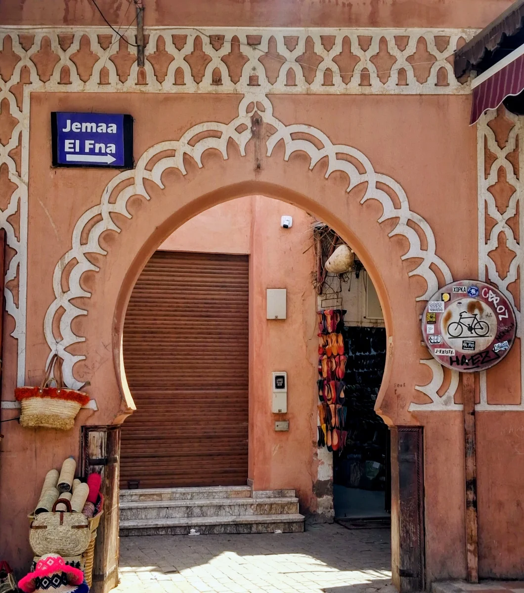 Traditioneller rosafarbener Lehmbogen, Wegweiser nach Jemaa el-Fna und Kunsthandwerksläden in einer belebten Straße des Tallâa-Viertels nahe der Medersa Ben Youssef.